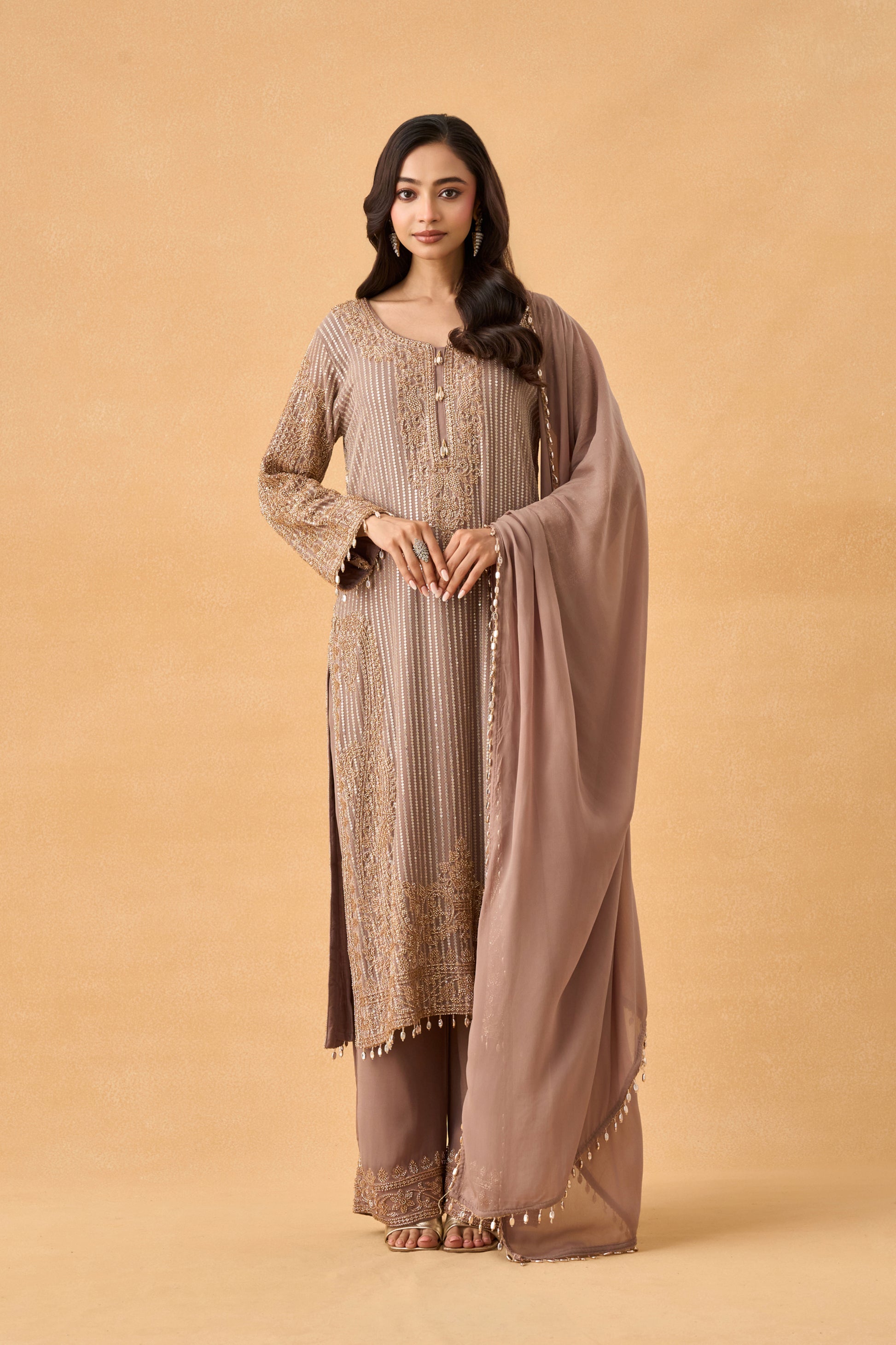 A woman wearing a traditional three-piece salwar kameez suit in taupe brown, featuring a long-sleeved embroidered kameez top, matching trousers, and a sheer dupatta, set against a solid beige studio background.