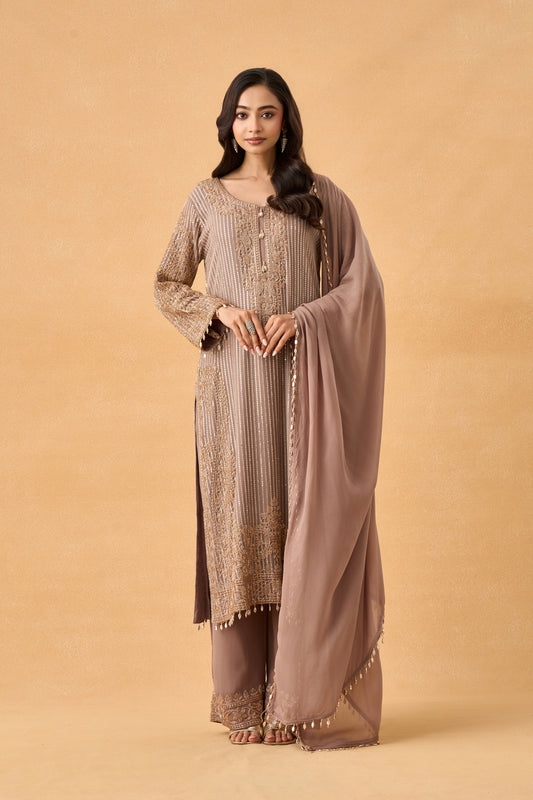 A woman wearing a traditional three-piece salwar kameez suit in taupe brown, featuring a long-sleeved embroidered kameez top, matching trousers, and a sheer dupatta, set against a solid beige studio background.