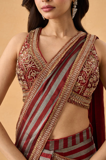 A close-up of a model wearing a traditional Indian draped saree featuring a red and gray striped saree with intricate gold embroidery and a matching embroidered sleeveless blouse.