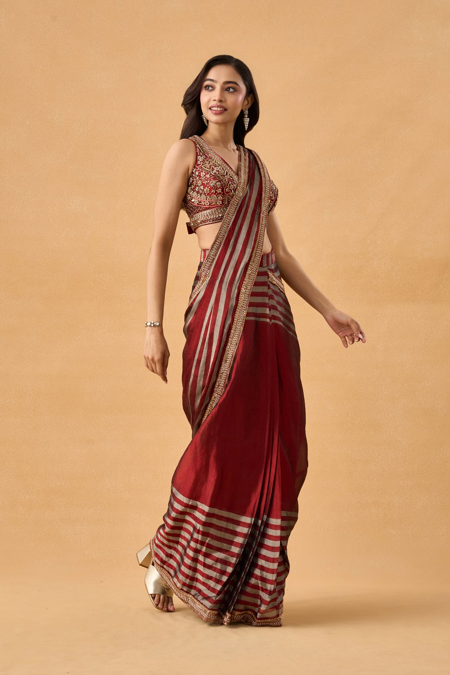 A woman wearing a traditional Indian pre draped saree with a deep red body, red and gold stripes, and a heavily embroidered gold and red blouse, set against a solid beige background.