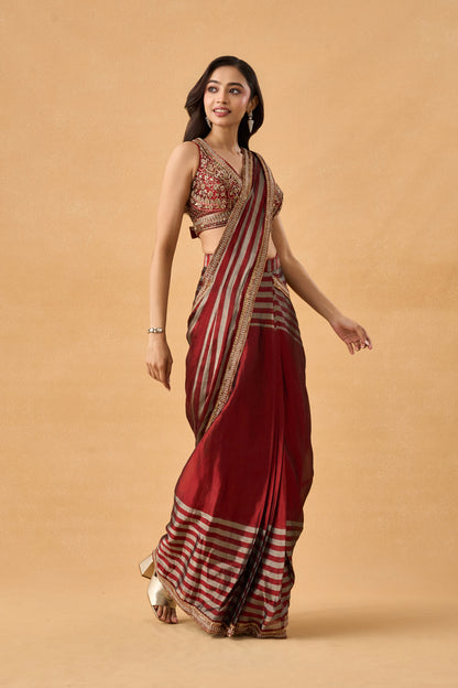 A woman wearing a traditional Indian pre draped saree with a deep red body, red and gold stripes, and a heavily embroidered gold and red blouse, set against a solid beige background.